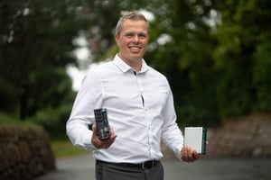 Mr. Gøran Labrå is holding two Moxa units, of which Hatteland Technology is distributor. There are green trees in the background.