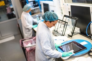 Two workers are performing optical bonding/preparations on marine monitors at Hatteland Technology's production facility. They are wearing lab suits and hair nets.