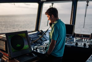 A marine navigational officer is standing in front of a HATTELAND® Series 1 monitor, while he is reporting by VHF. He is on a ship's bridge. You can see the ocean in the background, through the windows.